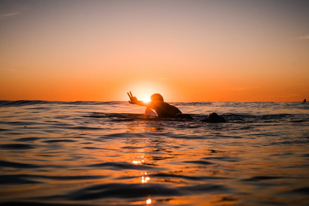 A surfer sits on their board at sunset, throwing a peace sign as the sun dips into the ocean